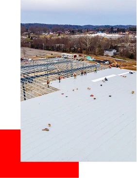 Aerial view of workers constructing a large metal building roof, with visible steel framework and scattered tools. Be Dry Roofing provides expert roofing services in Quincy IL. Trees, parked cars, and distant buildings are in the background.