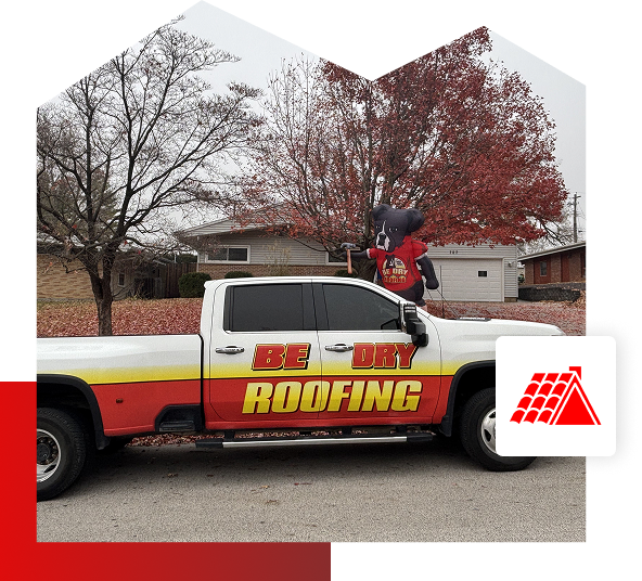 A white and red pickup truck with BE DRY ROOFING written on the side is parked on a residential street in Quincy IL. A large roof-themed mascot stands in the truck bed, with leafless trees and houses in the background.
